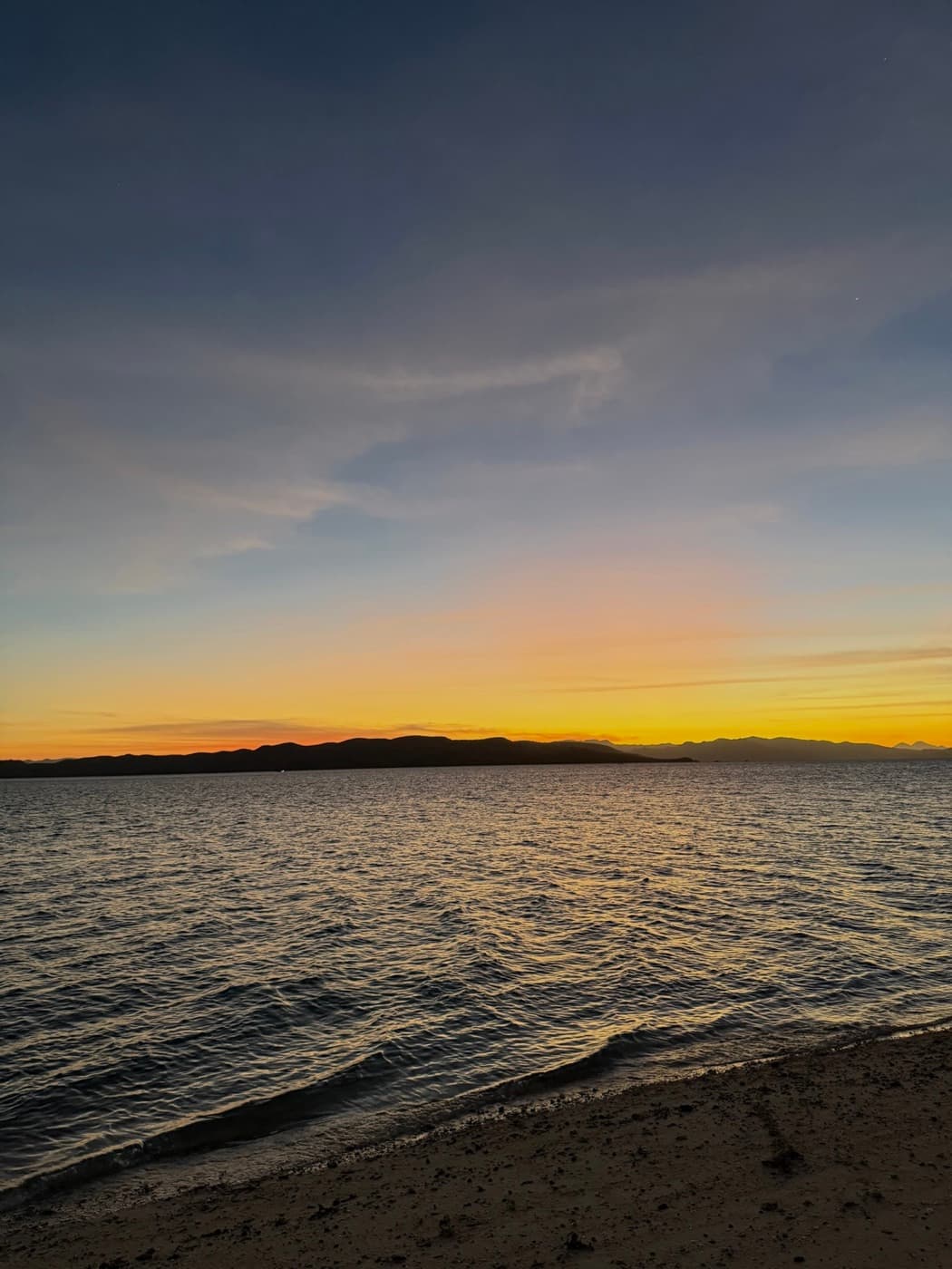 Sonnenuntergang über dem Meer, im Vordergrund der Strand, am Horizont eine Inselkette