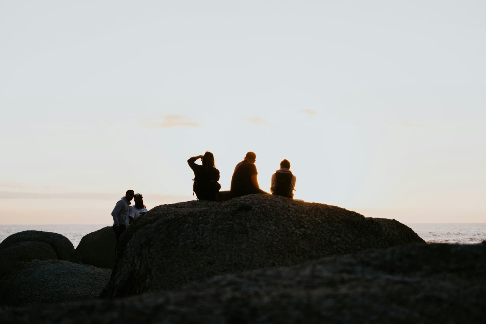 A small group sitting on rocks by the sea as silhouettes at sunset