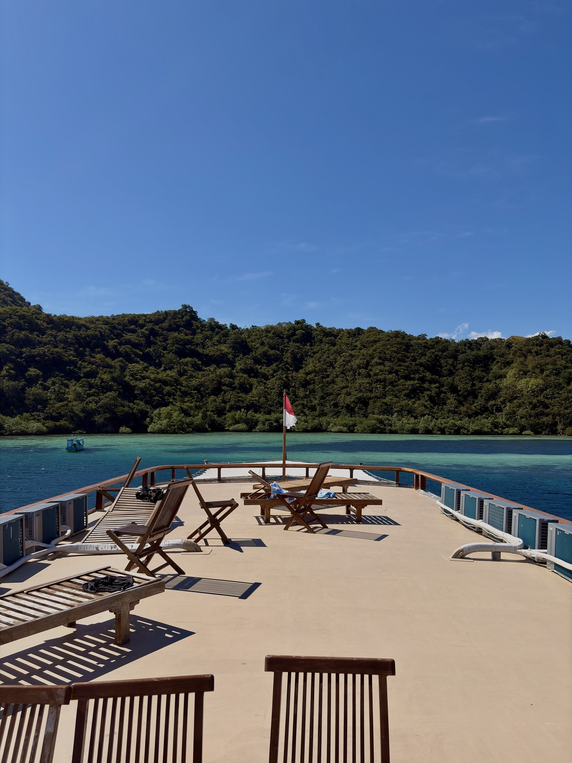 Holzliegen auf einer Terrasse über dem Wasser, dahinter bewaldete Hügel